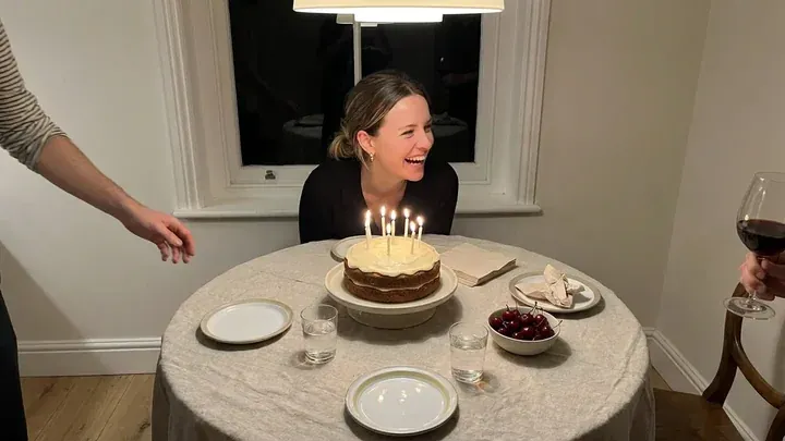 Nora laughing behind her birthday cake with the candles lit, friends reaching in at the edges